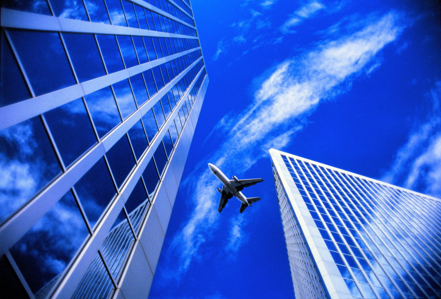 A low-angle shot captures two modern skyscrapers against a vibrant blue sky with wispy clouds. An airplane flies between the buildings, creating a sense of scale and movement within the urban landscape. The image evokes a feeling of travel and architectural wonder.