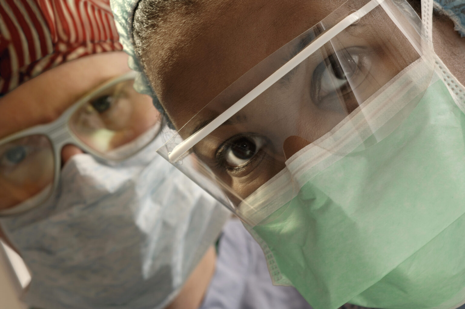Two healthcare workers in full PPE, including face masks and face shields, look intently towards the camera. The image highlights the protective gear worn by medical professionals during a pandemic, emphasizing safety and the importance of PPE. Photo by David Joel